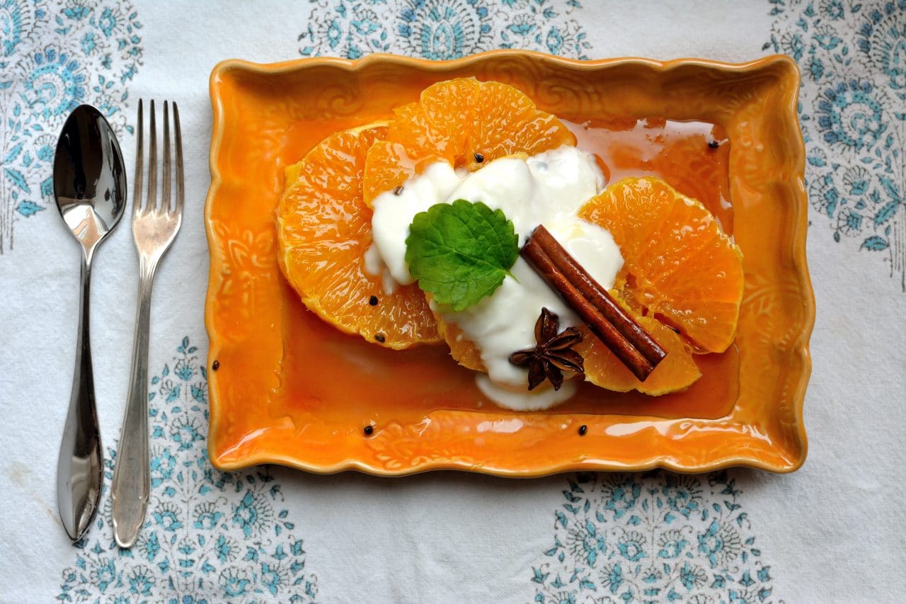 En orange tallrik med skivad apelsin med getmjölksyoghurt och två kanelstänger.