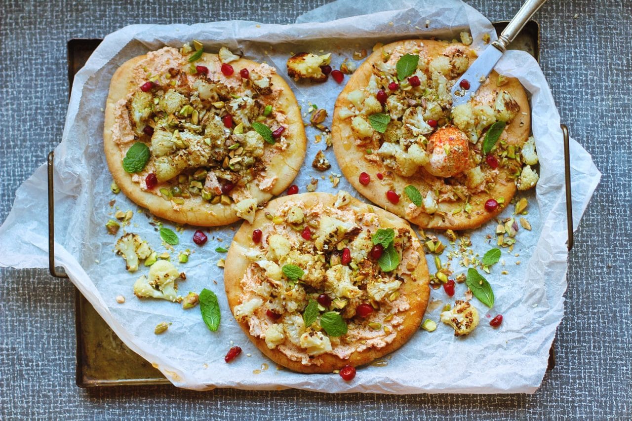 Tre runda pizzor på en bakplåt, pizzan är toppad med blomkål, labneh och granatäpplekärnor.