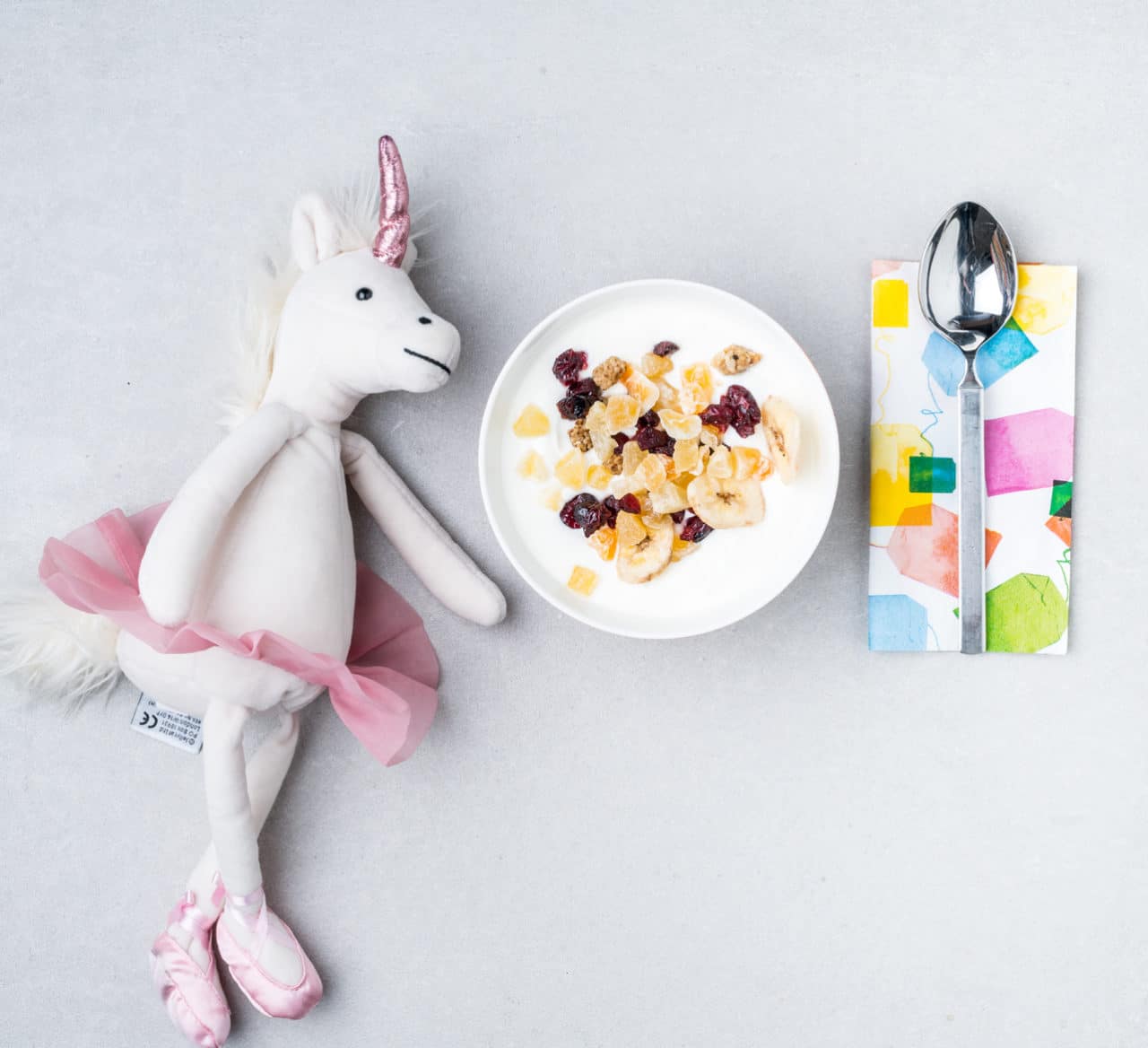 En vit och rosa leksak och ett mellanmål med Larsa yoghurt toppad med nötter och torkad frukt.