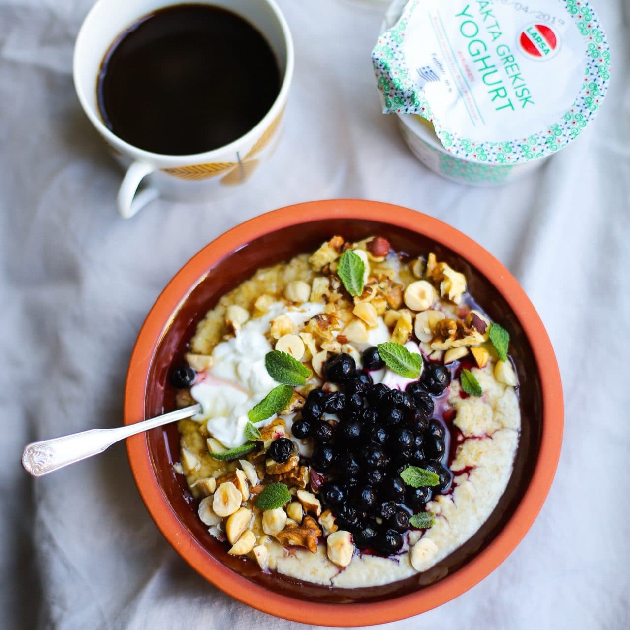 En kopp med svart kaffe och en skål med gröt toppad med Larsa yoghurt, blåbär, rostade hasselnötter och myntblad.